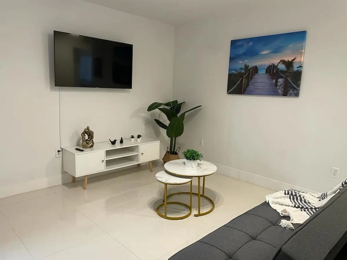 The hero image showcases a modern, inviting living room with a wall-mounted flat screen TV, sleek white TV stand adorned with decorative accents, a pair of chic marble-top coffee tables with gold frames, and a lush green potted plant in the corner. The space exudes comfort and style, perfect for relaxation after exploring Miami's vibrant Little Havana neighborhood.