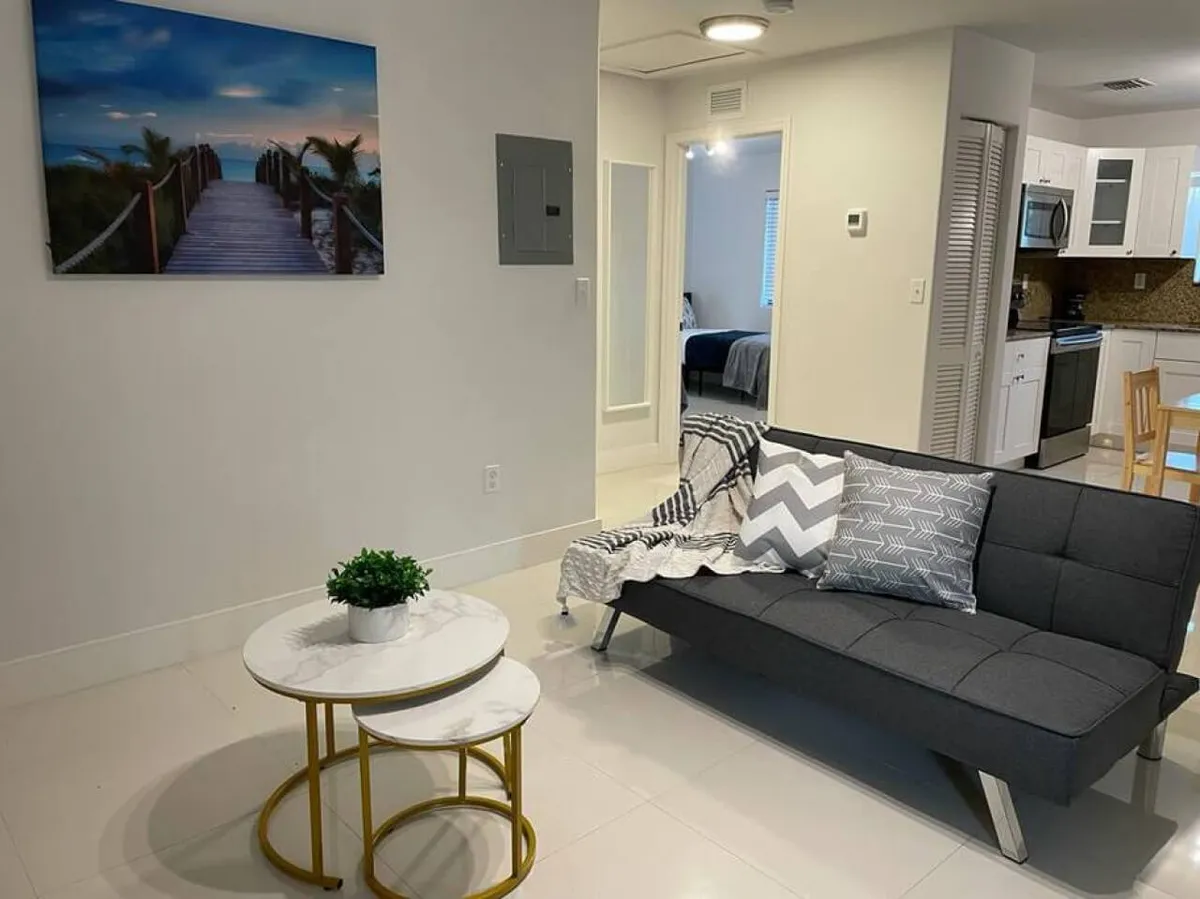 This image presents a bright and inviting open-plan living area featuring a dark gray sofa adorned with patterned pillows and a throw blanket. The space includes nesting round coffee tables, a small green plant, and an open kitchen area with modern appliances in the background. The living room is flooded with natural light, and the bedroom entrance is visible, adding depth to the space.