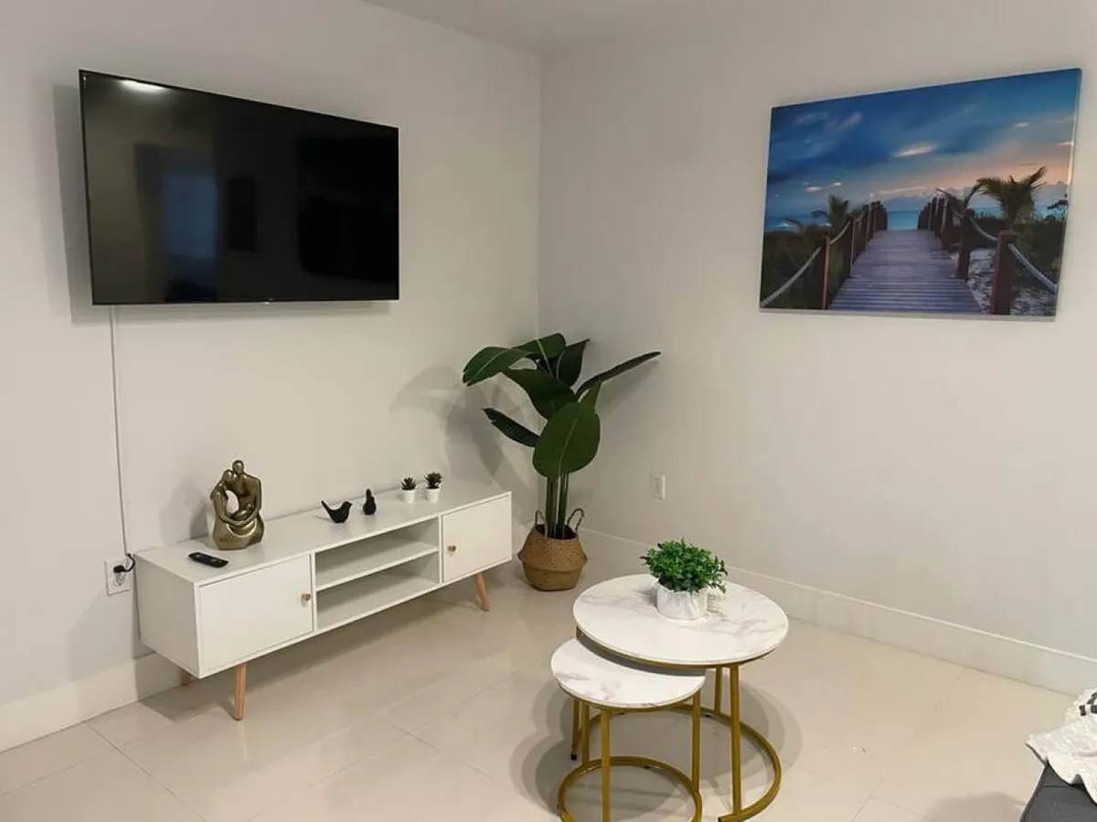 A peaceful and uncluttered living space centered around a wall-mounted flat screen TV and a white floating media console with decorative items. Nested round white marble tables with gold legs offer convenient surfaces, while a large green potted plant in the corner brings in a natural element. The decor is simple yet elegant, echoing Miami's stylish atmosphere.