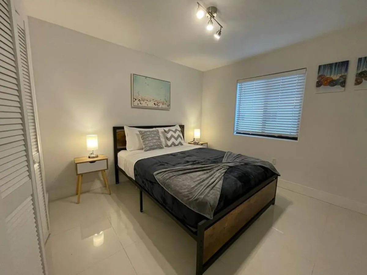 A softly-lit bedroom featuring a comfortable bed dressed in crisp linens and decorative pillows, flanked by two bedside tables with lamps. The window with blinds and the tasteful wall art above the bed add to the tranquil ambiance.