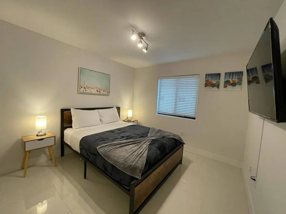 An inviting bedroom interior featuring a neatly made bed, two bedside tables with lamps, a wall-mounted flat-screen TV, and decorative artwork. The atmosphere is restful and private, ideal for unwinding after a day in Miami.