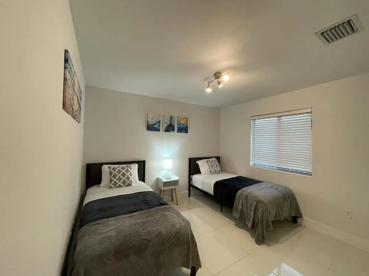 A minimalist bedroom with two twin beds featuring neutral bedding and dark frames. A small white nightstand with a lamp sits between the beds, and three-piece wall art and a window with closed blinds complete the uncluttered space.