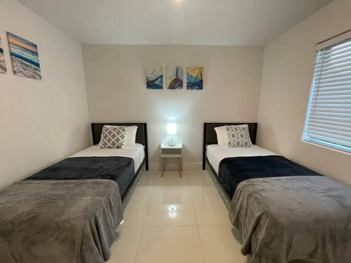 The image depicts a calm, restful bedroom with two twin beds featuring coordinated bedding and pillows. A small bedside table with a softly glowing lamp sits between the beds, while abstract artwork and a sunlit window create a cozy, inviting atmosphere.