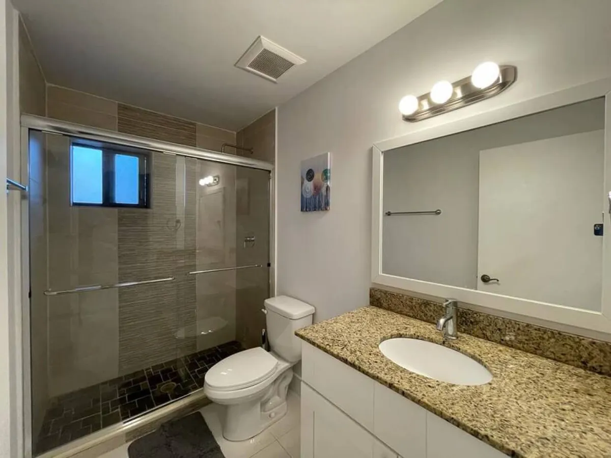 A clean, modern bathroom with a glass-enclosed shower, granite countertop sink, large framed mirror, and ample towels. The space is bright and organized, underscoring comfort and convenience.