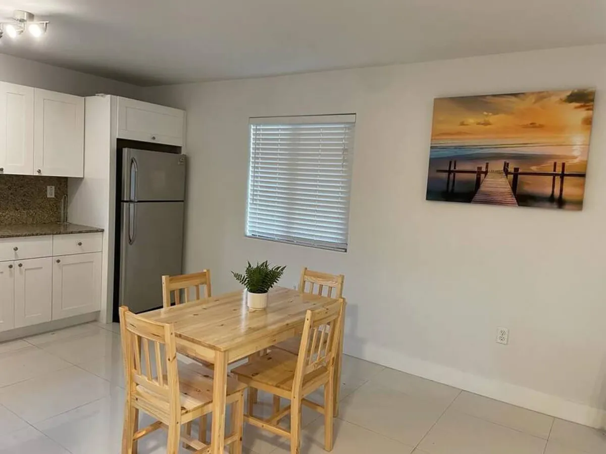 This image highlights a wooden dining table with four chairs set in a bright, inviting space. Behind the table, a modern stainless steel refrigerator and sleek kitchen cabinets are visible, along with a granite countertop. The scene is anchored by a large wall art depicting a tranquil sunset over a pier, adding both warmth and character to the room.