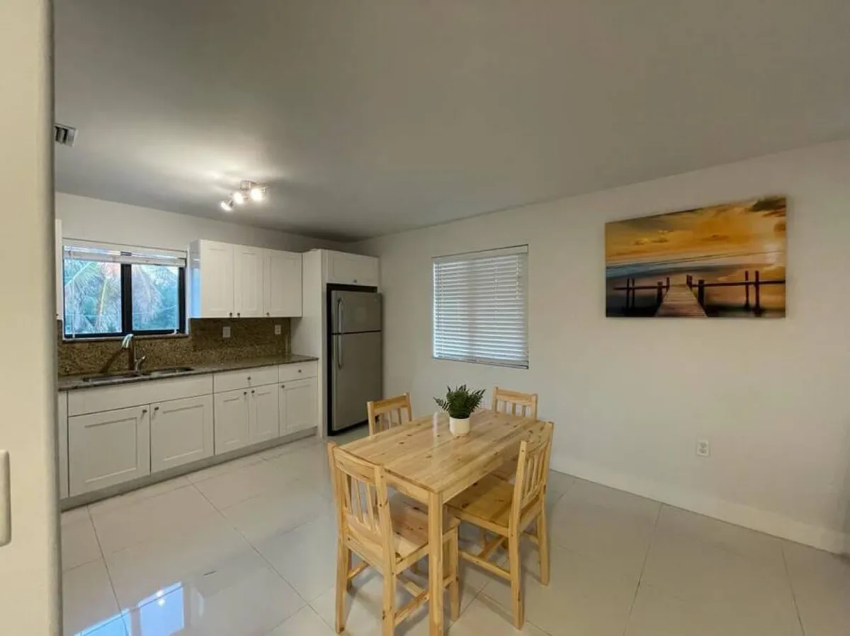 The image presents a bright, modern kitchen featuring white cabinets, granite countertops, and a stainless steel refrigerator. In the foreground is a wooden dining table with four chairs, set for casual meals or group conversations. The wall art of a sunset pier adds a touch of local flavor and ambiance to the space.