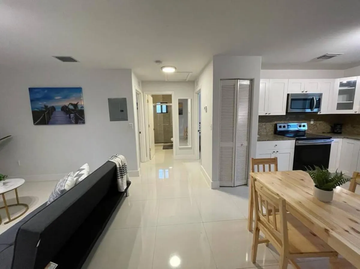 A spacious, welcoming open-plan living area with a black sofa adorned with cushions and a throw, a wooden dining table with chairs and a plant centerpiece, and a sleek white kitchen with stainless steel appliances. The hallway leads to additional rooms, suggesting a functional, social layout perfect for gatherings and relaxation.