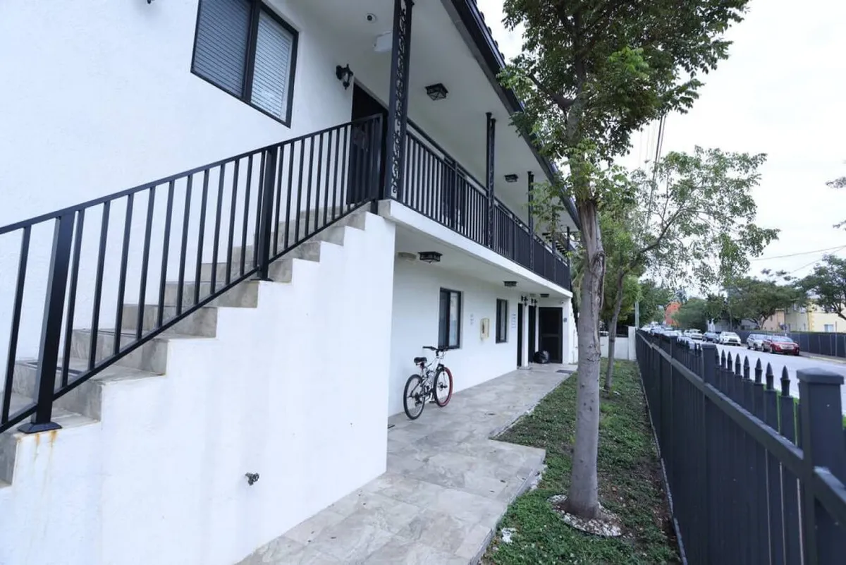This image shows the exterior of the building with a black-railed staircase leading up to the upper-level apartment. Two bicycles are parked by the wall, and a white building with black window frames is bordered by a fenced sidewalk lined with trees. The scene conveys both security and the convenience of direct access from parking to the apartment via stairs.